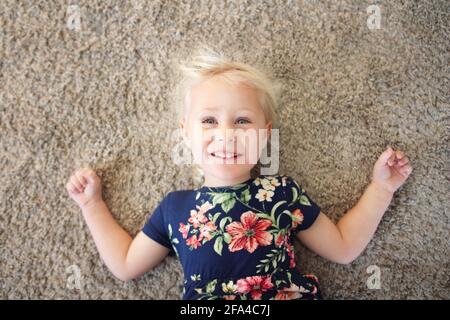 Una bambina felice in un vestito di fiore sta posando sul suo tappeto di casa sorridente. Foto Stock