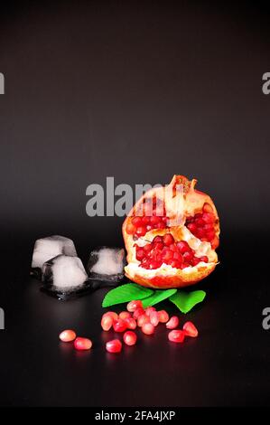Un melograno, un frutto maturo con foglie spezzate e adagiate su uno sfondo nero accanto a semi e cubetti di ghiaccio fuso. Primo piano. Foto Stock