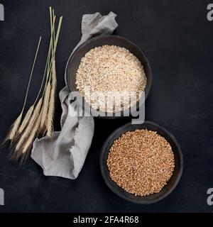 Fiocchi di avena rotolati in ciotole nere e orecchie di grano dorato su sfondo scuro. Vista dall'alto Foto Stock