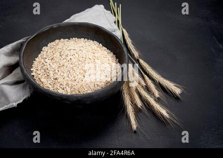 Fiocchi di avena rotolati in ciotole nere e orecchie di grano dorato su sfondo scuro. Foto Stock