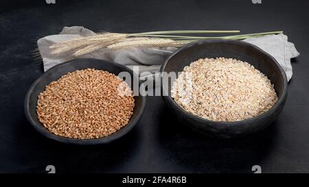 Fiocchi di avena rotolati in ciotole nere e orecchie di grano dorato su sfondo scuro. Foto Stock