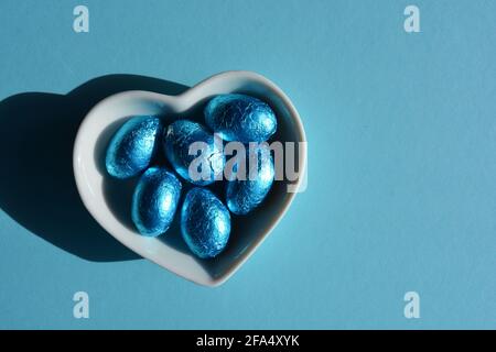 Uova di Pasqua al cioccolato avvolte con lamina blu in un piatto a forma di cuore isolato sullo sfondo Foto Stock