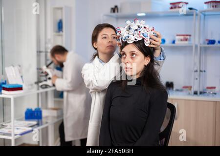 Donna che indossa cuffie a onde cerebrali in un moderno laboratorio di medicina con medico neurologo. Neuroscience medic che mette i sensori sul paziente. Trovare la cura per la malattia. Foto Stock