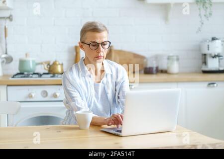 Moderna donna specializzata scrittore maturo indossare occhiali digitando qualcosa sul computer portatile, lavorando online mentre si siede in cucina a casa. Concetto di persone anziane e tecnologie Foto Stock