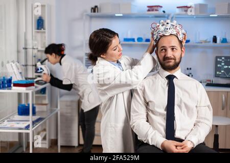 Donna ricercatrice che ha messo le cuffie per la scansione delle onde cerebrali al paziente sviluppando esperimenti utilizzando i monitor di laboratorio per lo studio cerebrale. Uomo che si siede sulla sedia mentre lo scienziato che regola il dispositivo che esamina il sistema nervoso Foto Stock