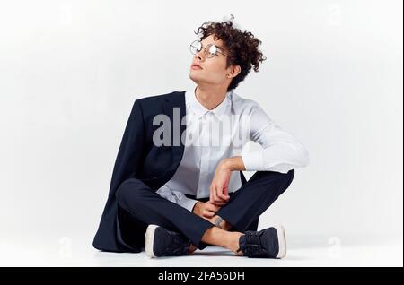 un ragazzo dai capelli ricci in abiti e sneaker alla moda si siede sul pavimento in una stanza luminosa in piena crescita Foto Stock