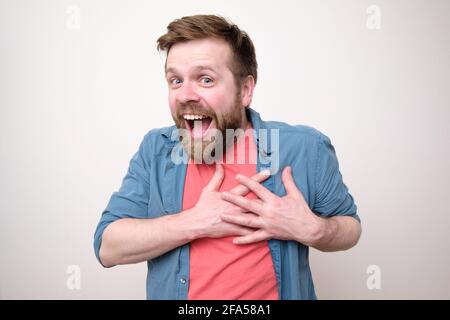 Gioioso uomo caucasico è sorpreso di una piacevole sorpresa, sorride ampiamente, tiene le mani sul cuore e guarda la fotocamera. Foto Stock