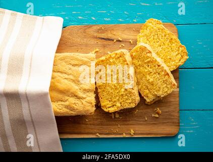 Il pane di mais è un pane contenente farina di mais e farina di mais. Bel pane di mais fatto in casa giallo affettato sotto un asciugamano di cotone. Sfondo di legno blu. Foto Stock