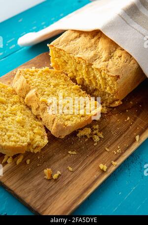 Il pane di mais è un pane contenente farina di mais e farina di mais. Bel pane di mais fatto in casa giallo affettato sotto un asciugamano di cotone. Sfondo di legno blu. Foto Stock