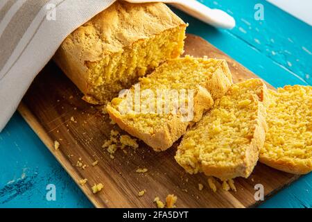 Il pane di mais è un pane contenente farina di mais e farina di mais. Bel pane di mais fatto in casa giallo affettato sotto un asciugamano di cotone. Sfondo di legno blu. Foto Stock