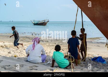 Attività di pesca sulla spiaggia di Shannah, Oman. Uomo e ragazzi seduti all'ombra dell'arco di un dhow Foto Stock