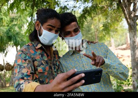 Giovani amici maschi indiani in maschere di protezione medica viso prendendo un selfie che mostra la vittoria sig Foto Stock