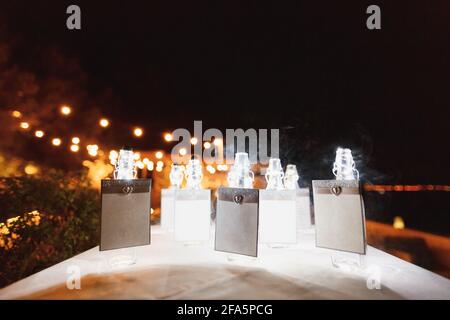 I flaconi sigillati con cartelli in bianco si trovano su un tavolo coperto con una tovaglia bianca sullo sfondo di una luce brillante lampadine di notte Foto Stock