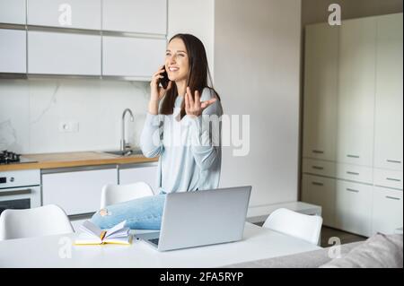 Giovane donna affascinante che usa il computer portatile per lavorare a distanza da casa, parla sullo smartphone appoggiato sul tavolo in posizione rilassata, freelance femminile ha una piacevole conversazione telefonica Foto Stock