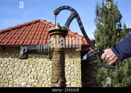 La mano dell'uomo tiene la maniglia di un vecchio rubinetto dell'acqua di strada. Sullo sfondo e' un edificio in pietra con tetto in tegole rosse. Stile di vita rustico. Primo piano. Messa a fuoco selettiva. Foto Stock