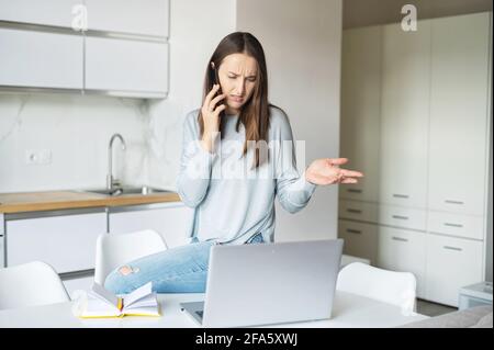 Infastidita giovane donna parla sullo smartphone utilizzando un computer portatile per lavorare da casa, irritata donna guarda lo schermo del portatile con conversazione telefonica Foto Stock