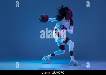 Bella african-american giocatore di basket femminile in movimento e in azione in luce al neon su sfondo blu. Concetto di stile di vita sano, professionale Foto Stock