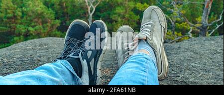 banner con immagine concettuale di due coppie di gambe in sneakers sullo sfondo della roccia forestale. Scarpe da ginnastica unisex casual e jeans in montagna. Cou Foto Stock