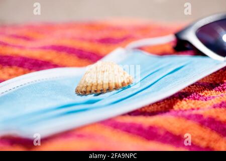 primo piano di una maschera blu monouso, con una conchiglia in cima, accanto ad un paio di occhiali da sole su un asciugamano arancione, sulla spiaggia Foto Stock