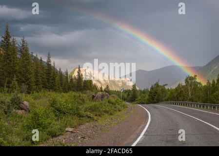 Incredibile arcobaleno su un cielo scuro e tempestoso con nuvole su una tortuosa strada asfaltata, montagne e foreste. Altai, Russia Foto Stock