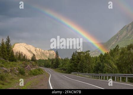 Incredibile arcobaleno su un cielo scuro e tempestoso con nuvole su una tortuosa strada asfaltata, montagne e foreste. Altai, Russia Foto Stock