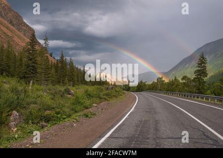 Incredibile arcobaleno su un cielo scuro e tempestoso con nuvole su una tortuosa strada asfaltata, montagne e foreste. Altai, Russia Foto Stock