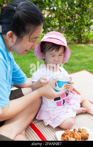 Vacanza, madre e adorabile ragazza asiatica (tailandese) godere di mangiare il loro pranzo nel parco, bambino che tiene cucchiaio e tazza, pollo fritto sul piatto, oltre Foto Stock