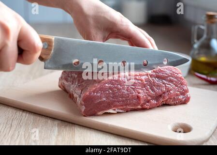 Le mani femminili tagliano un pezzo crudo di carne di vitello con un coltello in cucina, filetto su un tagliere, cucina a casa, primo piano. Foto Stock