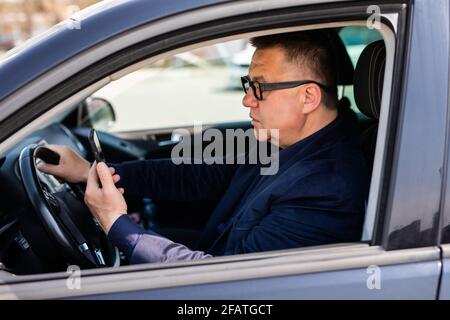 Un uomo d'affari di media età seduto utilizza un telefono cellulare in un'auto di guida. Foto Stock