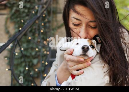 Attraente hipster giovane donna in occhiali da sole baciare jack russell terrier cucciolo nel parco, prato verde e fogliame sfondo. Divertente cane purebred baciato da Foto Stock