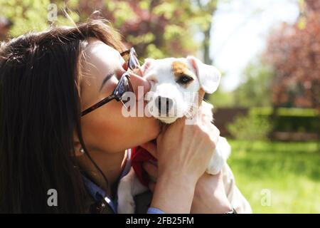 Attraente hipster giovane donna in occhiali da sole baciare jack russell terrier cucciolo nel parco, prato verde e fogliame sfondo. Divertente cane purebred baciato da Foto Stock