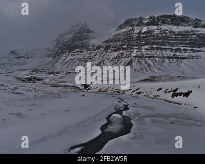 Paesaggio aspro con fiume ghiacciato e montagne ripide innevate con ripide pendii vicino a Grundarfjörður sulla penisola di Snæfellsnes, Islanda occidentale. Foto Stock