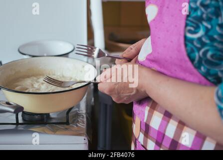 Donna di mezza età che cucina con forcelle in mani. Ciotola di pasta su stufa Foto Stock