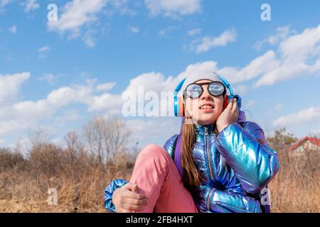 Adolescente ragazza in occhiali da sole specchiati guarda nel cielo. Concetto di benessere. Il bambino ascolta musica e canta Foto Stock