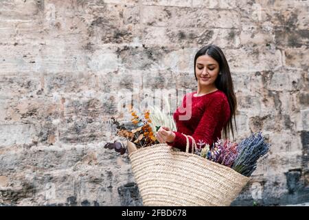 Bella donna che trasporta sacchetto di paglia di fiori mentre sta in piedi a. parete Foto Stock