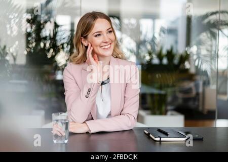 Felice imprenditrice che guarda lontano mentre è seduto alla scrivania in ufficio Foto Stock