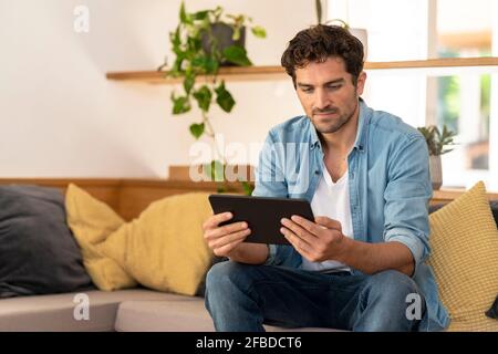 Uomo serio che usa il tablet digitale mentre si siede sul divano a. casa Foto Stock