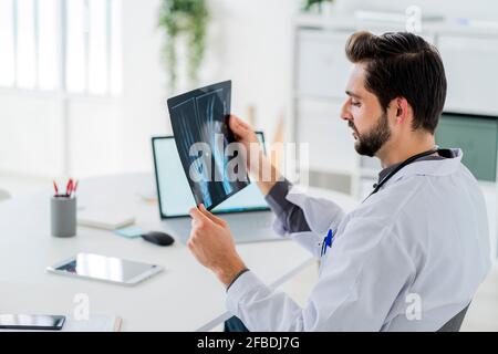 Lavoratore sanitario di sesso maschile che analizza i raggi X mentre si siede alla scrivania ospedale Foto Stock