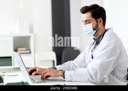 Medico di sesso maschile indossando la maschera mentre si usa il computer portatile alla scrivania Foto Stock