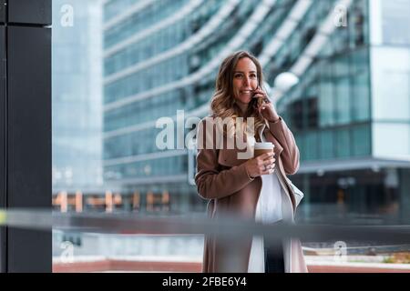 Donna d'affari sorridente con i capelli biondi che hanno caffè mentre parla sopra telefono cellulare Foto Stock