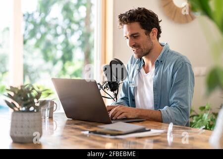 Freelance maschile che parla sul microfono di fronte al computer portatile mentre registrazione di podcast a casa Foto Stock