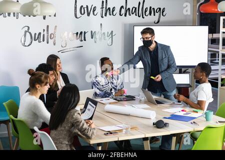 Uomo imprenditore indossando maschera protettiva che dà nota adesiva a. colleghi in riunione Foto Stock