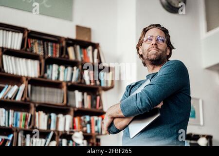 Proprietario maschile maturo con braccia incrociate e portatile da libreria presso la caffetteria Foto Stock