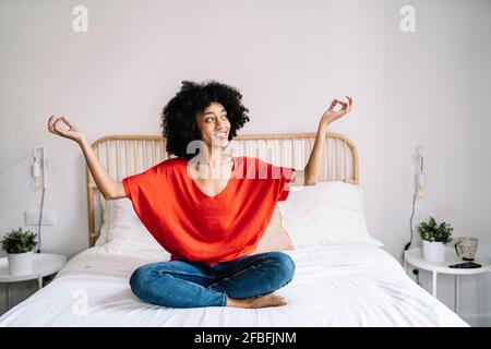 Sorridente giovane donna meditante mentre si siede a letto a casa Foto Stock