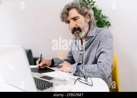 Professionista maschile senior che guarda il notebook mentre utilizza la grafica tablet a casa Foto Stock