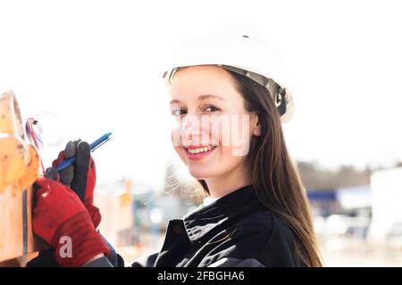 Sorridente ingegnere femminile che scrive sulla clipboard in cantiere Foto Stock