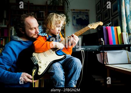 Padre che insegna chitarra elettrica al figlio biondo mentre si siede a. casa Foto Stock