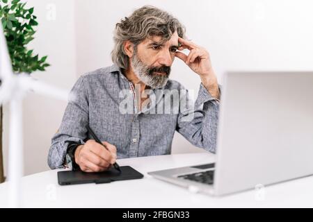 Un uomo d'affari senior concentrato con un tablet grafico che guarda il notebook mentre seduto in ufficio Foto Stock