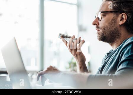 Imprenditore maschile che invia la posta vocale tramite smartphone tramite laptop in caffè Foto Stock