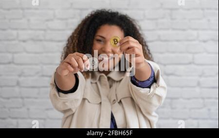 Donna sorridente che mostra bitcoins color argento e oro davanti al muro Foto Stock